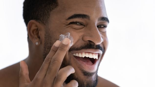 Homem sorridente com pele hidratada e de aspecto saudável aplicando protetor solar facial com toque seco no rosto com os dedos.