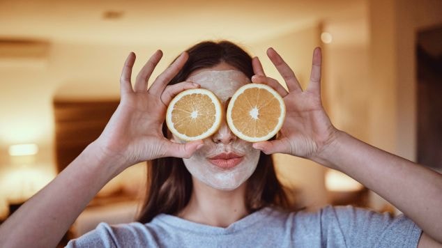 Mulher sorridente fazendo skincare com vitamina C segurando duas fatias de laranja na frente dos olhos.