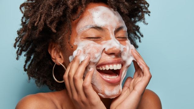 Mulher sorridente com pele de tipo oleosa lavando o rosto com as mãos com sabonete facial líquido, com o rosto e mãos cobertos de espuma.
