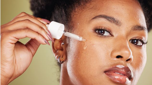 Mulher negra com os cabelos presos em um coque aplicando sérum no rosto.