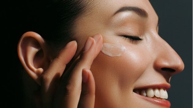 Mulher branca sorrindo aplicando com as mãos um creme de textura leve com vitamina C, deixando sua pele hidratada e radiante.