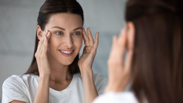 Mulher com pele uniforme e bem cuidada, sorriso leve e expressão tranquila, tocando a pele após aplicar um hidratante facial com toque seco.