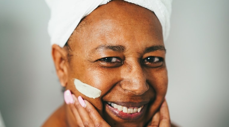 Senhora negra sorri, enquanto utiliza creme com ação antissinais em sua pele facial.
