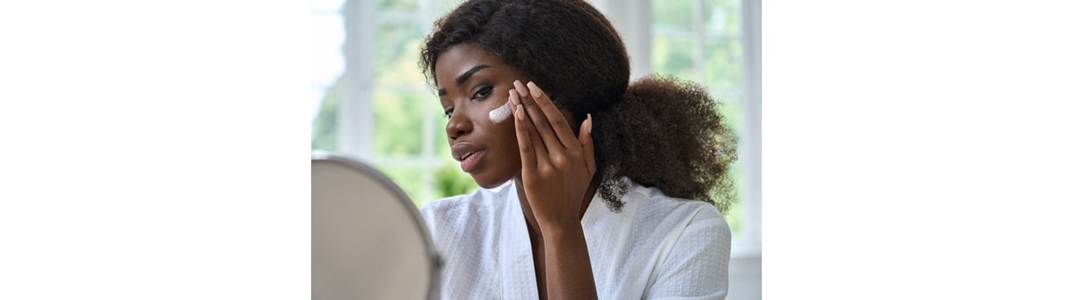 Moça negra, em frente ao espelho, aplica hidratante com nicotinamida em sua pele do rosto