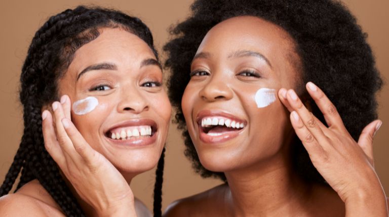 Duas jovens mulheres negras sorridentes aplicam produtos da rotina skincare na pele do rosto