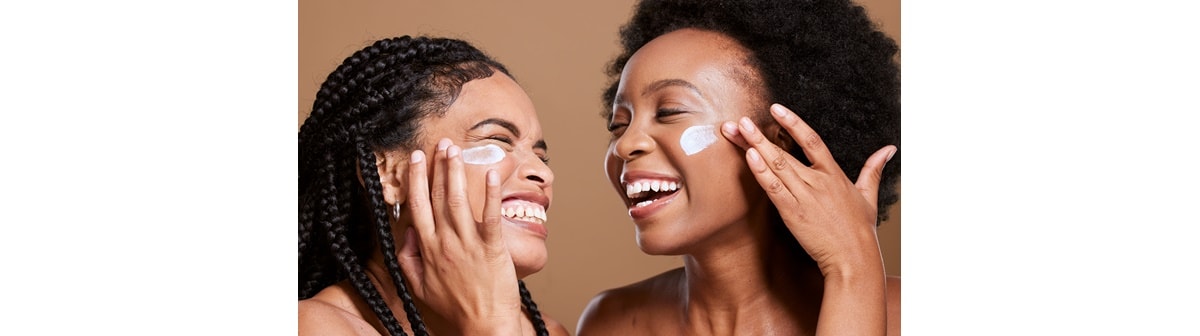 Duas mulheres pretas sorrindo e com o olho fechado passando hidratante