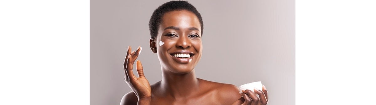 Jovem mulher negra sorri ao hidratar seu rosto que pode ter pele oleosa