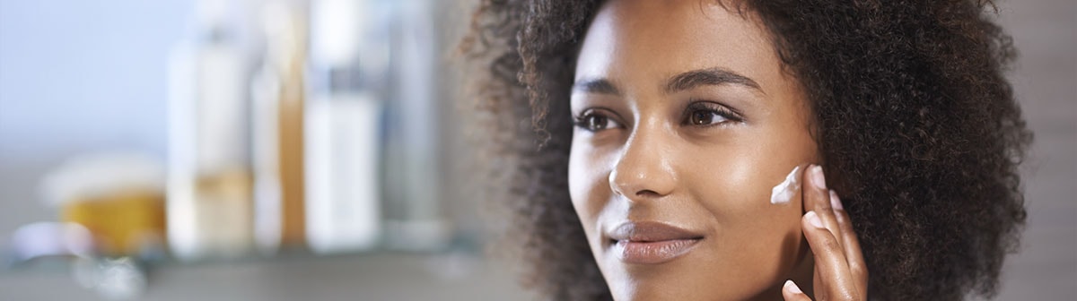 mulher negra com postura séria aplicando creme facial na pele do rosto
