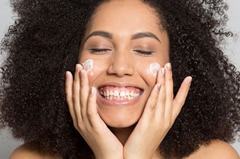 mulher negra sorridente aplicando creme nos dois lados do rosto