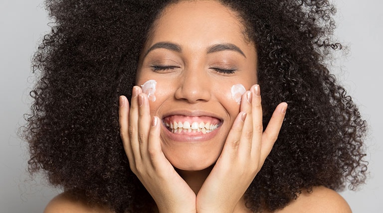 mulher negra sorridente aplicando creme nos dois lados do rosto