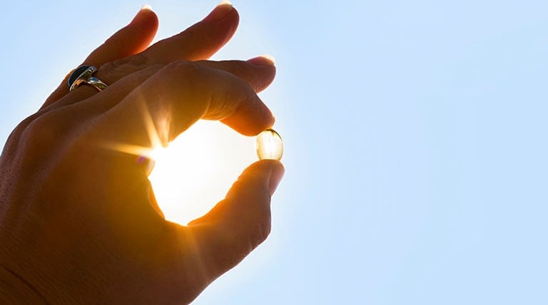 Mão segurando uma cápsula de vitamina do sol e luz solar ao fundo