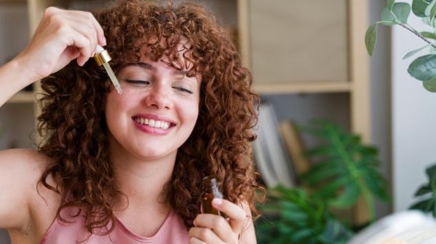 Jovem branca e ruiva com o cabelo cacheado aplica um sérum facial com alfa-arbutin para reduzir manchas no rosto