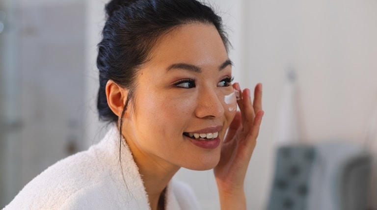 Mulher asiática sorri ao aplicar creme com colágeno e vitamina c no rosto para rejuvenescer.