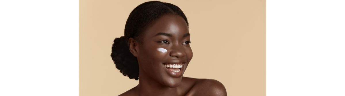 Jovem negra sorridente utiliza creme para hidratar o rosto