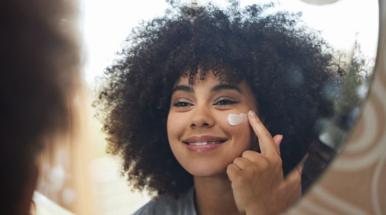Jovem sorridente com o cabelo estilo black power aplica o protetor solar na pele do rosto em frente ao espelho