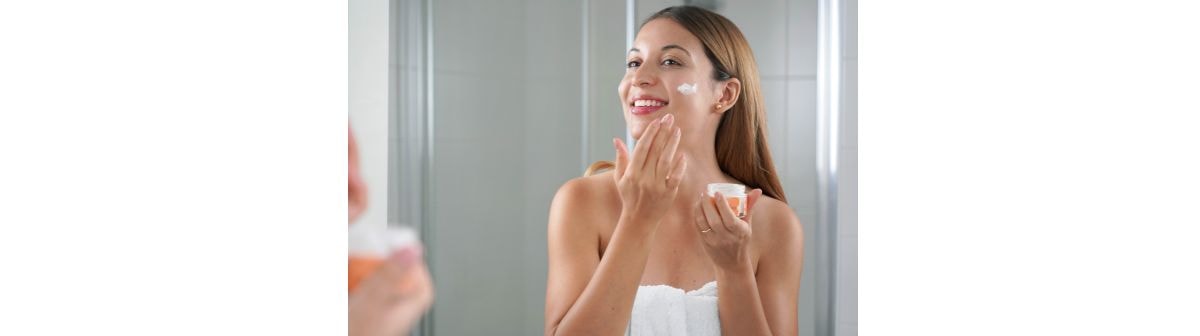Mulher sorrindo em frente ao espelho enquanto aplica creme  no rosto, representando rotina de skincare para redução de manchas e uniformização da pele