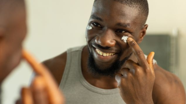 Homem sorrindo aplicando creme de rosto na região dos olhos, refletido no espelho, em sua rotina de skincare.