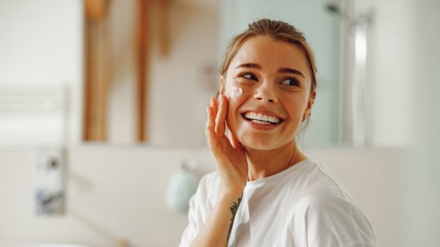 Jovem mulher sorridente aplica um creme facial em seu rosto