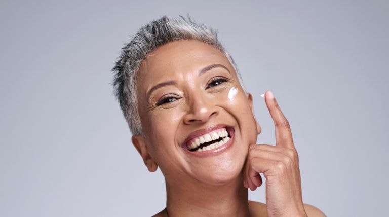 Mulher de pele madura sorri ao utilizar um creme hidratante em seu rosto
