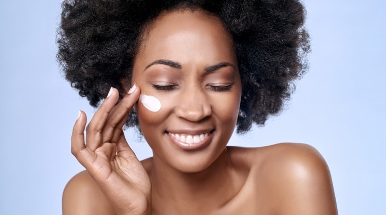 Mulher negra aplica creme hidratante do rosto ao sorrir de olhos fechados