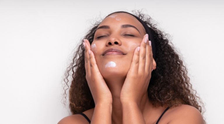 Jovem negra aplica creme hidratante no rosto para garantir a hidratação facial