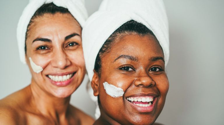Duas mulheres de etnias diferentes sorriem ao utilizar o creme hidratante para o rosto
