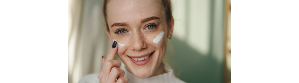 Jovem mulher ruiva sorri enquanto aplica um creme hidratante em sua pele do rosto