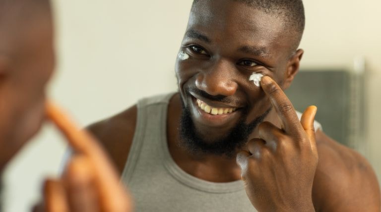 Homem negro sorri ao aplicar o creme hidratante para aproveitar seus benefícios