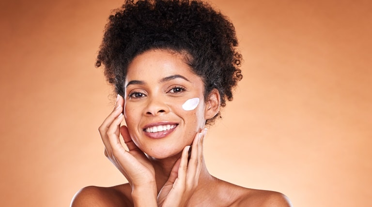 Mulher negra sorri ao aplicar creme hidratante para o rosto em sua pele facial