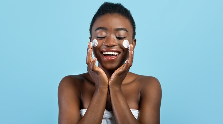 Jovem negra sorri ao aplicar creme no rosto em ambas as bochechas