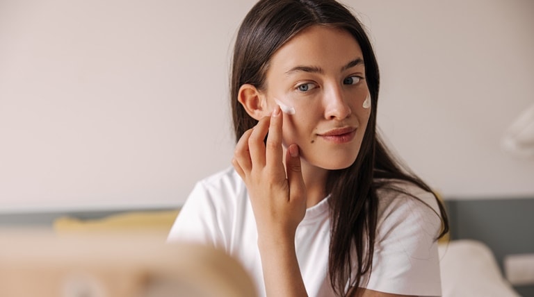 Jovem caucasiana aplica um hidratante para pele acneica no rosto em frente ao espelho.