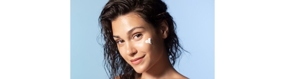 Jovem mulher de cabelos cacheados sorri com uma porção de hidratante para pele oleosa em seu rosto, formando um coração