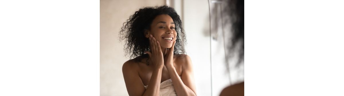 Jovem mulher negra e sorridente aplicando hidratante para pele sensível em seu rosto