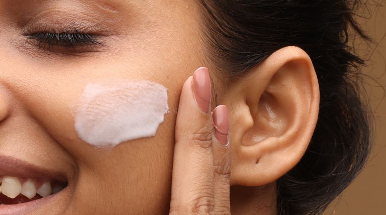 Mulher sorrindo enquanto aplica creme hidratante na bochecha, fazendo uma rotina de skincare.