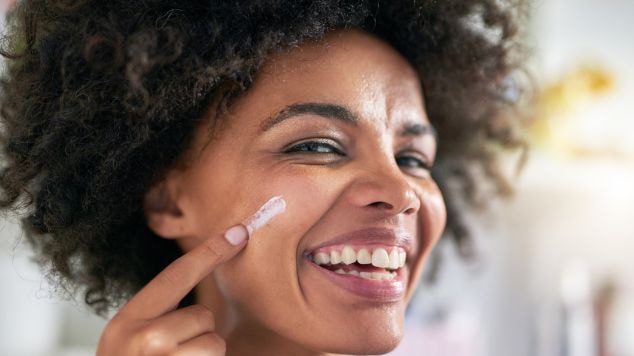Mulher sorridente aplicando vitamina C em creme diretamente no rosto.