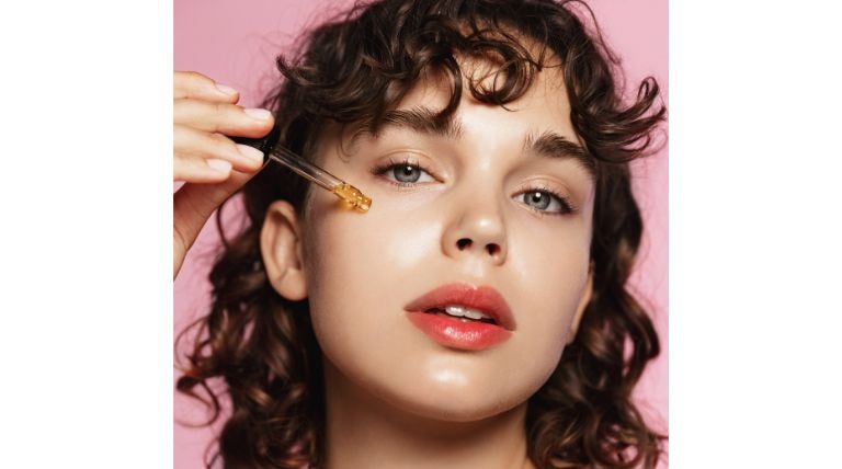 Mulher com cabelo cacheado aplicando um sérum facial com conta-gotas, parte da rotina de cuidados com vitamina C.