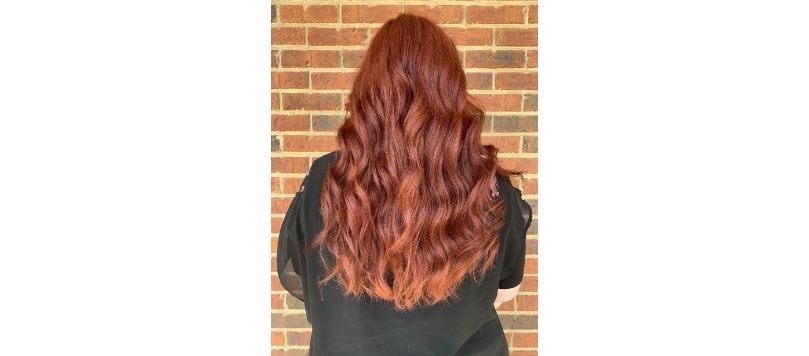 Mulher de costas com cabelo longo e ondulado em tons de cabelo vermelho escuro acobreado, com reflexos vibrantes que vão do vermelho ao cobre.