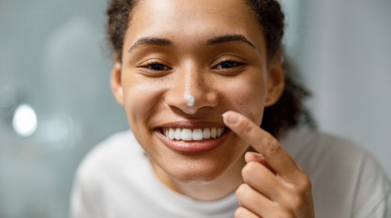 Mulher negra está sorridente ao aplicar um hidratante facial com toque seco em sua pele do rosto