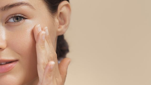 Mulher jovem com pele iluminada aplicando hidratante com aloe vera no rosto, olhos verdes e fundo bege neutro.