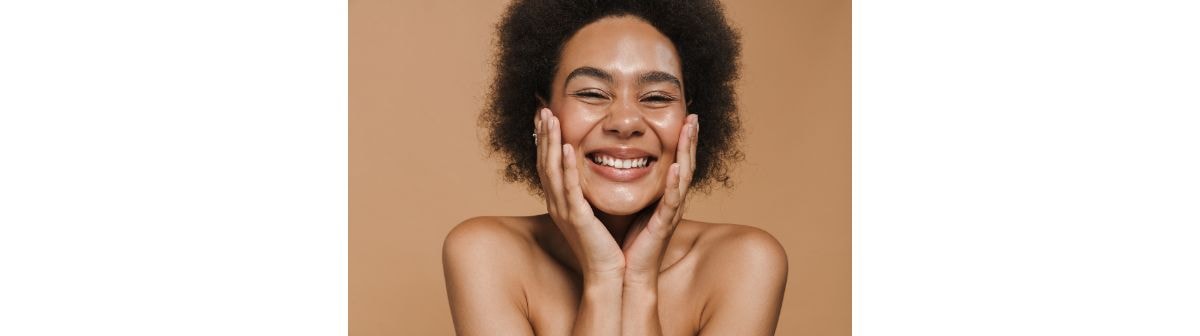 Mulher com cabelo cacheado sorrindo e segurando o rosto com as mãos, expressando felicidade com uma pele saudável.