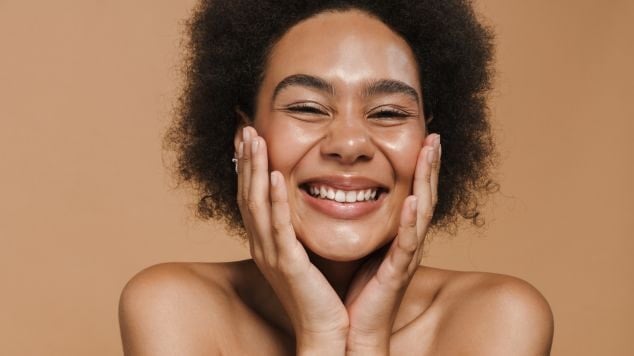 Mulher com cabelo cacheado sorrindo e segurando o rosto com as mãos, expressando felicidade com uma pele saudável.