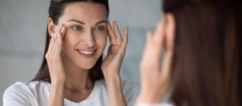 Mulher sorridente se olhando no espelho e tocando suavemente a região ao redor dos olhos com as pontas dos dedos.