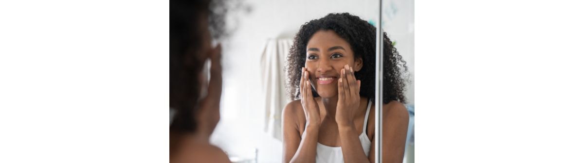 Uma mulher jovem de cabelos escuros e cacheados, sorri enquanto aplica um produto na bochecha com as duas mãos.