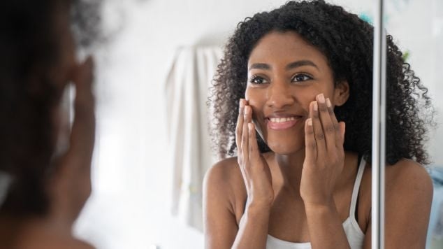 Uma mulher jovem de cabelos escuros e cacheados, sorri enquanto aplica um produto na bochecha com as duas mãos.