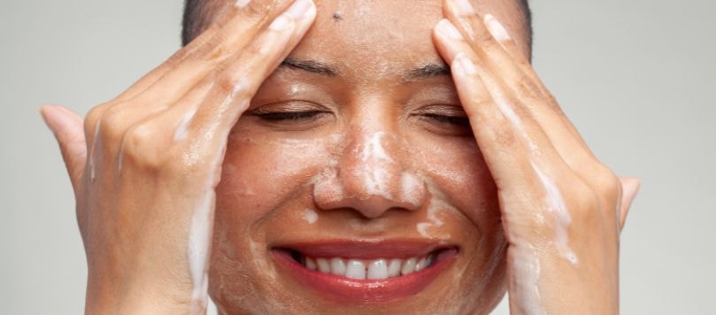 Mulher negra jovem sorrindo com olhos fechados lavando o rosto com espuma de limpeza facial para reduzir poros dilatados, fundo cinza.