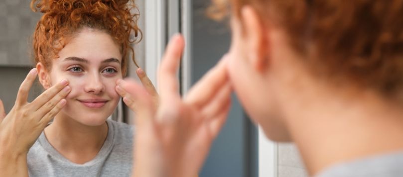 Mulher ruiva se olhando no espelho enquanto aplica hidratante facial.