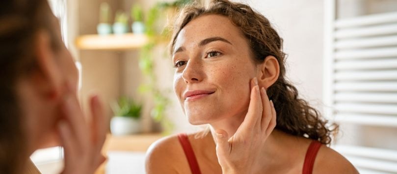 Mulher jovem com sardas tocando o rosto enquanto se olha no espelho em banheiro iluminado, representando rotina de skincare diária.