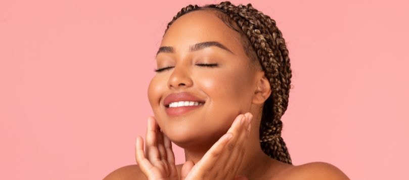 Mulher com pele morena e tranças com mechas claras, sorrindo com os olhos fechados e as mãos próximas ao rosto, sobre fundo rosa, evidenciando uma pele com aspecto hidratado e uniforme.