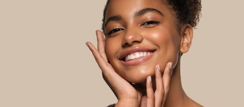 Mulher jovem de cabelos cacheados sorri com as mãos no rosto, exibindo pele luminosa e saudável, transmitindo confiança em sua rotina de skincare.