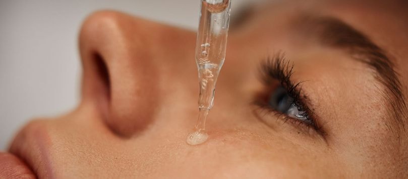 Aplicação de sérum facial com pipeta na bochecha de mulher de olhos azuis, close no rosto com pele clara e fundo cinza claro.